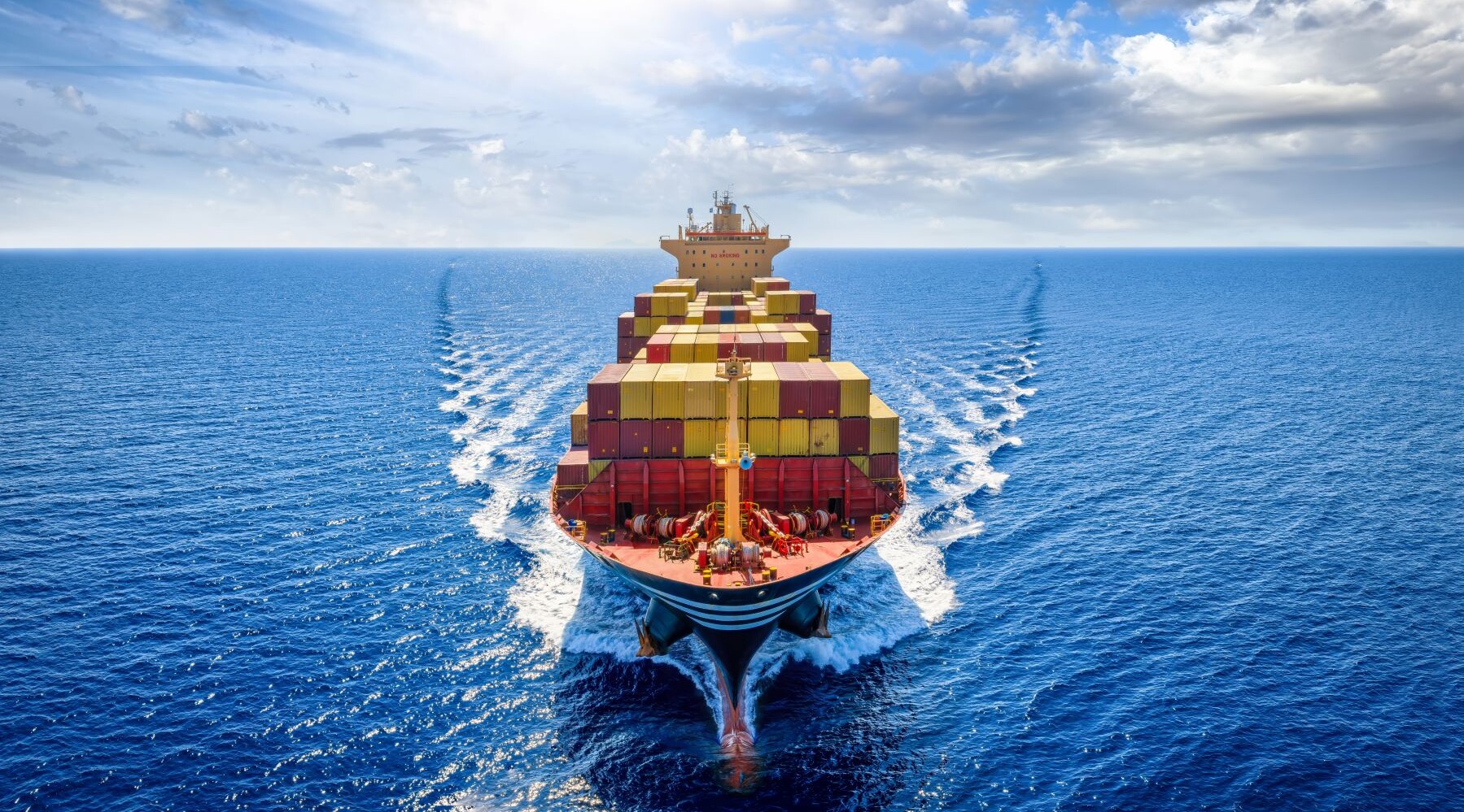 Containerschiff auf offener See, beladen mit gestapelten Frachtcontainern, fährt geradeaus durch ruhiges blaues Wasser unter einem teils bewölkten Himmel.