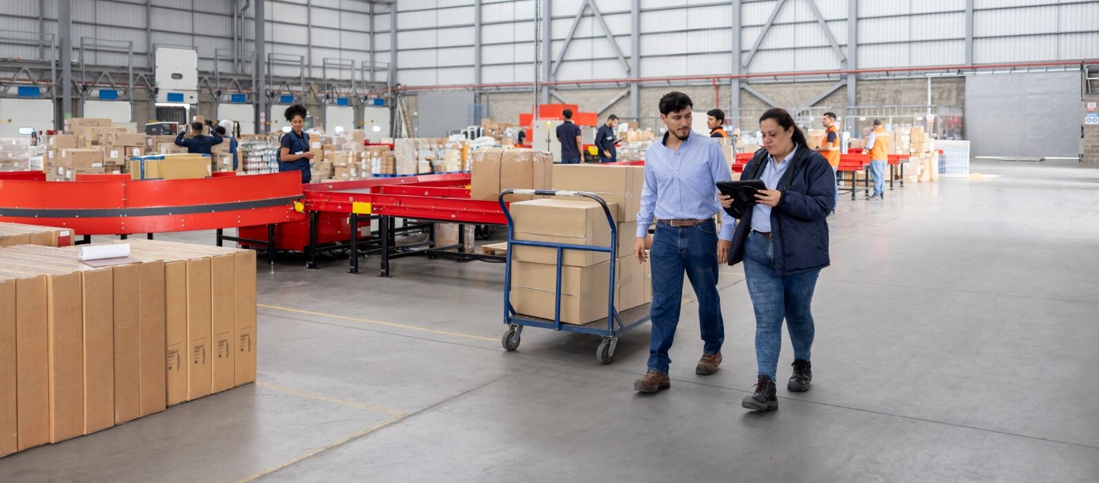 Zwei Mitarbeitende besprechen Logistikprozesse in einem Lagerhaus mit Paketwagen und Förderbändern im Hintergrund