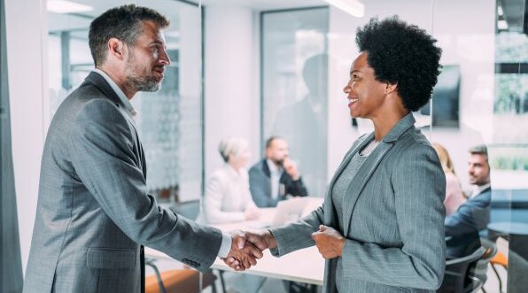 Zwei Geschäftspartner geben sich in einem modernen Büro die Hand, während ein Team im Hintergrund an einem Meeting teilnimmt.