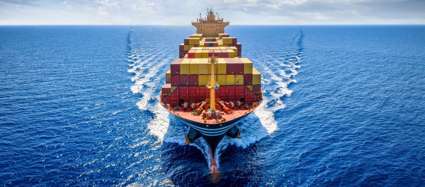 Containerschiff auf offener See, beladen mit gestapelten Frachtcontainern, fährt geradeaus durch ruhiges blaues Wasser unter einem teils bewölkten Himmel.