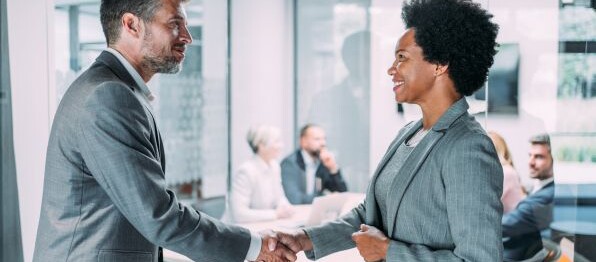 Zwei Geschäftspartner geben sich in einem modernen Büro die Hand, während ein Team im Hintergrund an einem Meeting teilnimmt.