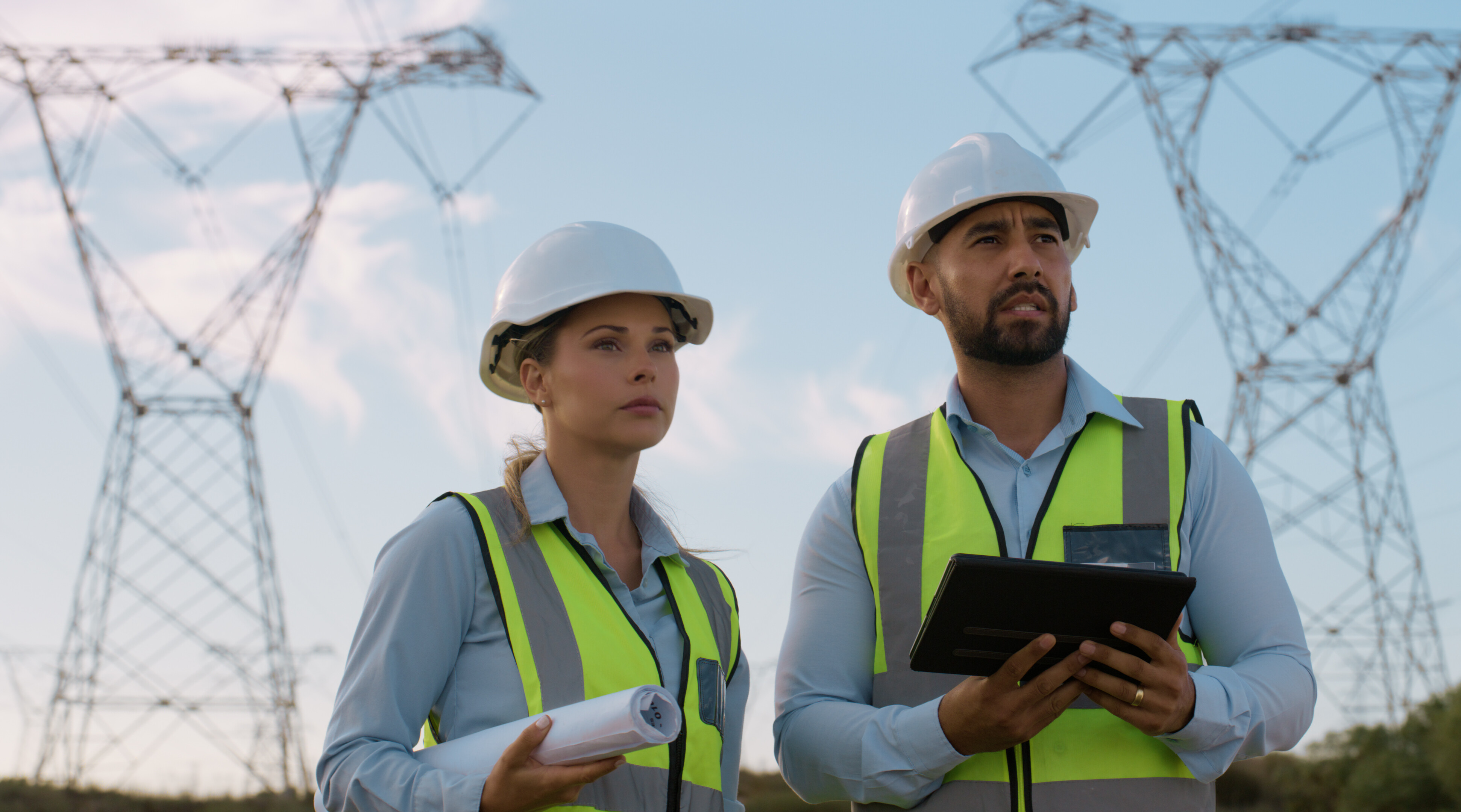 Zwei Ingenieure mit Schutzhelmen und Warnwesten stehen im Freien vor Hochspannungsmasten, halten Baupläne und ein Tablet und inspizieren eine Stromtrasse.
