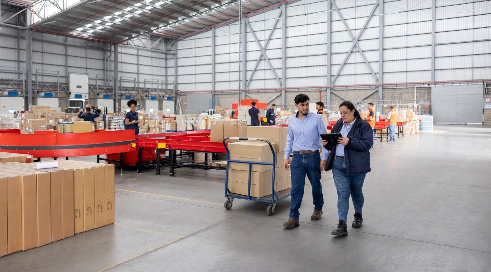 Zwei Mitarbeitende besprechen Logistikprozesse in einem Lagerhaus mit Paketwagen und Förderbändern im Hintergrund