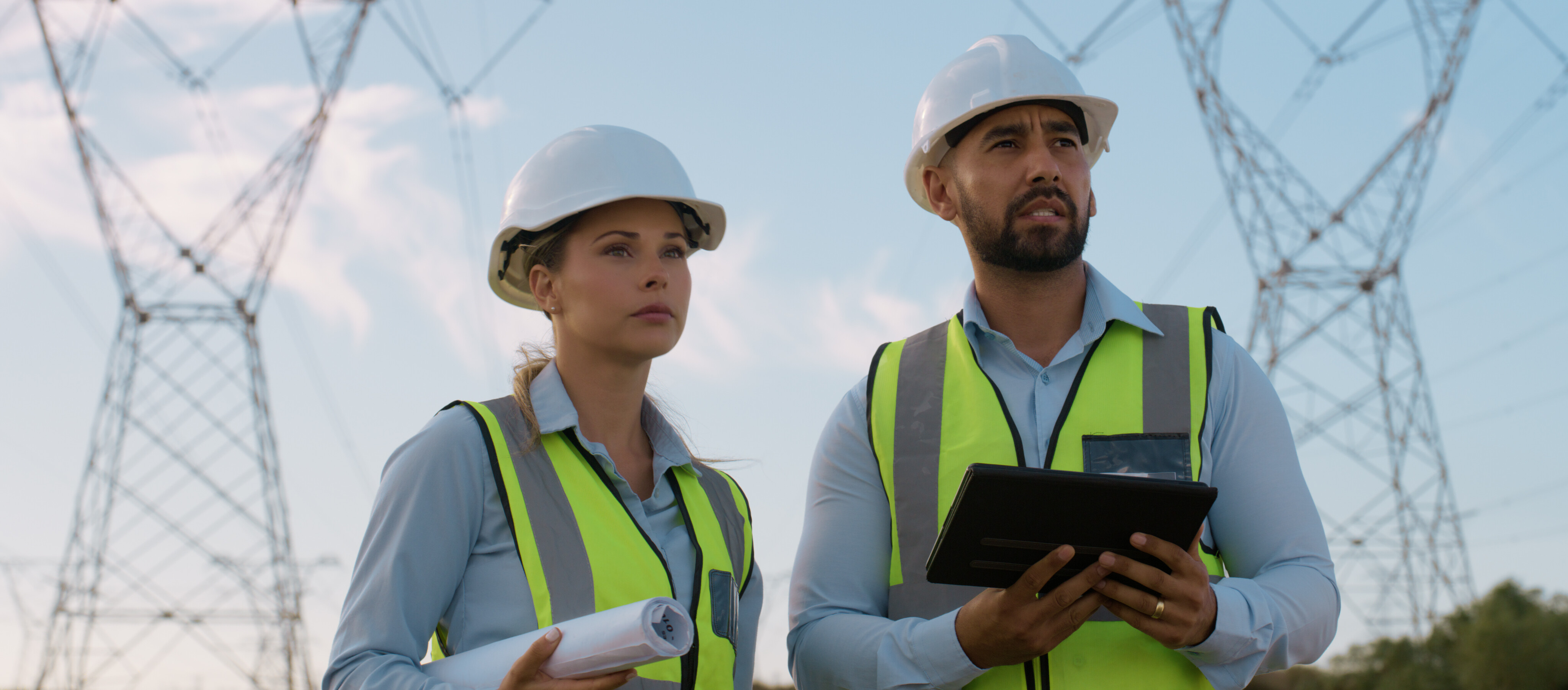 Zwei Ingenieure mit Schutzhelmen und Warnwesten stehen im Freien vor Hochspannungsmasten, halten Baupläne und ein Tablet und inspizieren eine Stromtrasse.