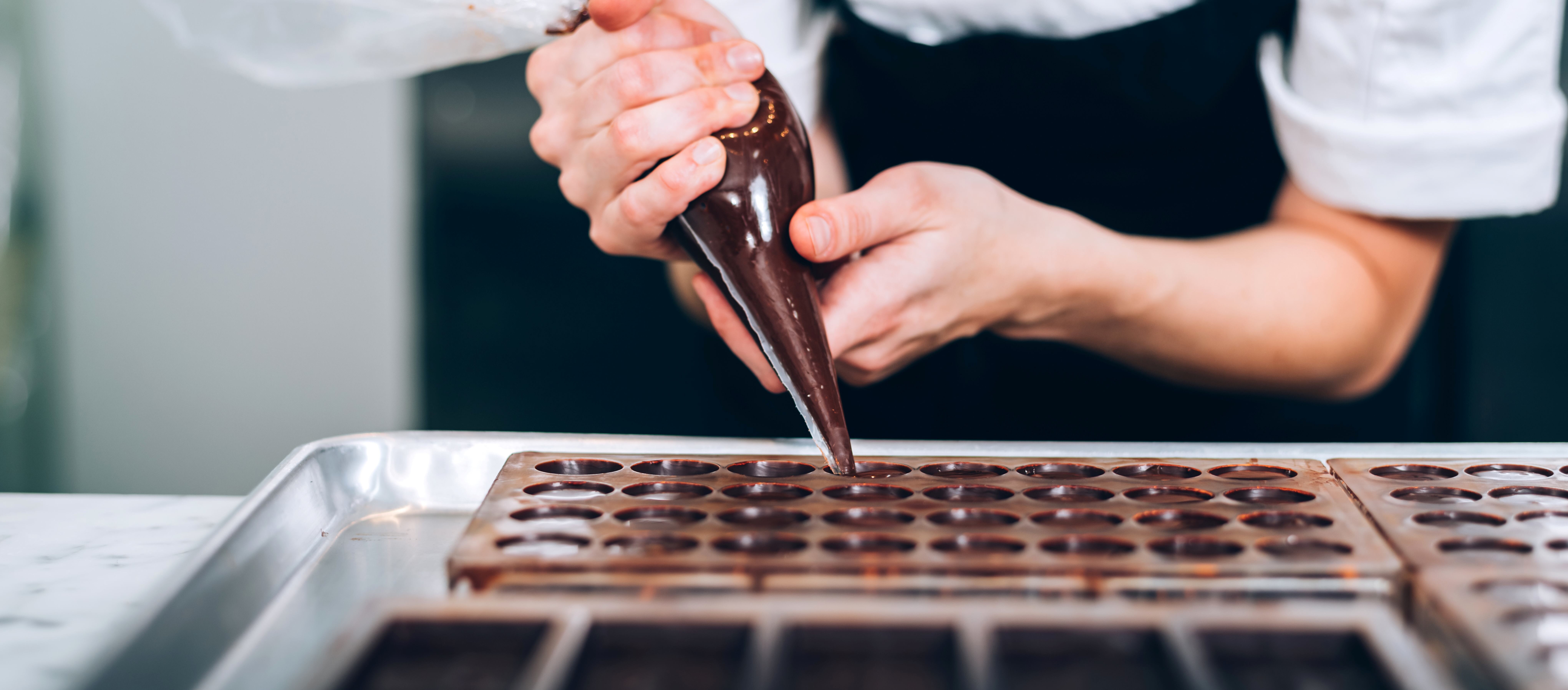 „Ein*e Chocolatier füllt mit einem Spritzbeutel vegane Schokolade in eine Form. Hände und Arbeitsutensilien stehen im Fokus.“